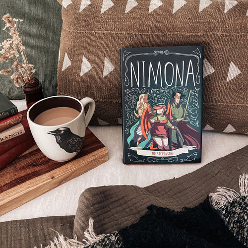 Illumicrate edition of Nimona by ND Stevenson leaning against a brown cushion, next to a mug of coffee and a book stack. image
